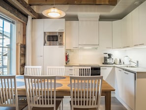 Una cocina moderna con armarios blancos, horno empotrado, microondas y cafetera. Hay una mesa de comedor con sillas y una lámpara colgante arriba.