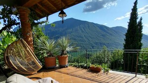 Une terrasse avec vue sur une montagne, une chaise en osier, des plantes en pot et une table avec une bouteille et des verres.