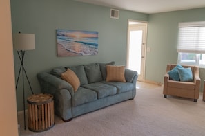 Un soggiorno con un divano grigio, un tavolino rotondo in legno, una lampada da terra, una poltrona beige e un quadro raffigurante una spiaggia.