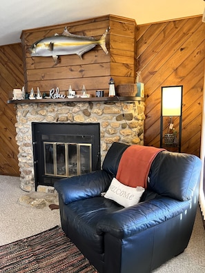Uma sala de estar acolhedora com lareira de pedra, uma poltrona de couro e um travesseiro com a inscrição 'Welcome', além de um peixe decorativo sobre a lareira.