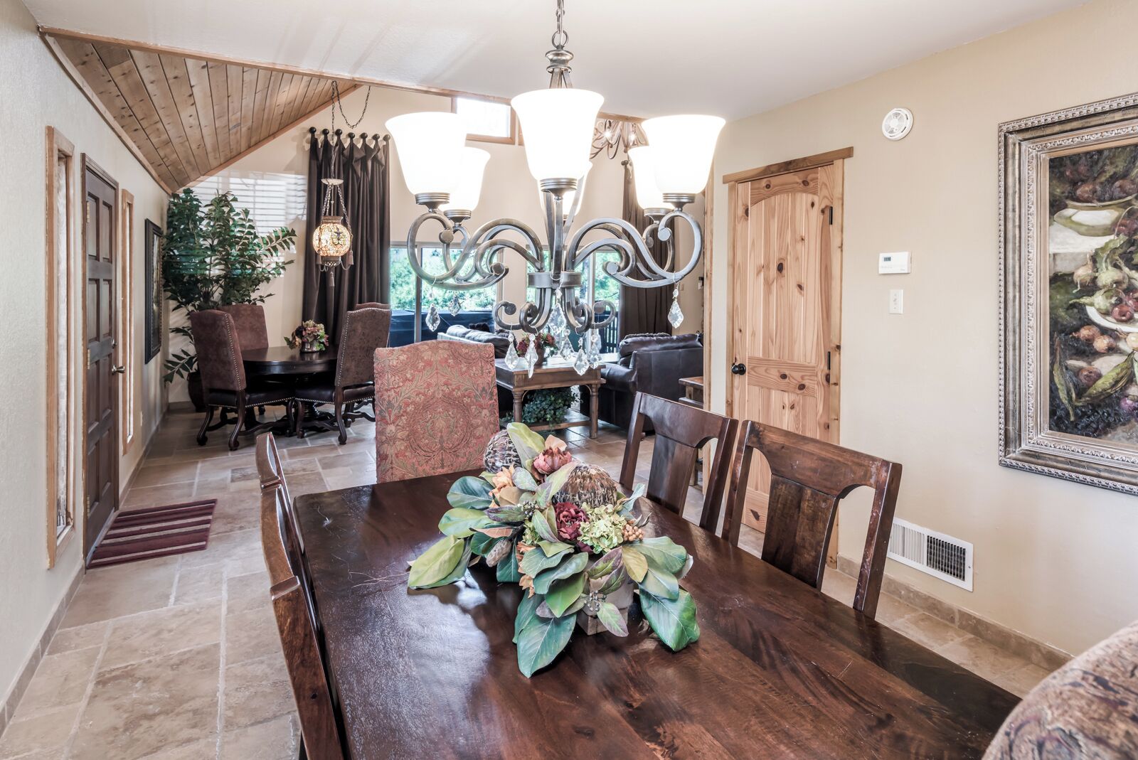 A dining area with a wooden table, chairs, and a chandelier. There is a potted plant on the table and a painting on the wall.
