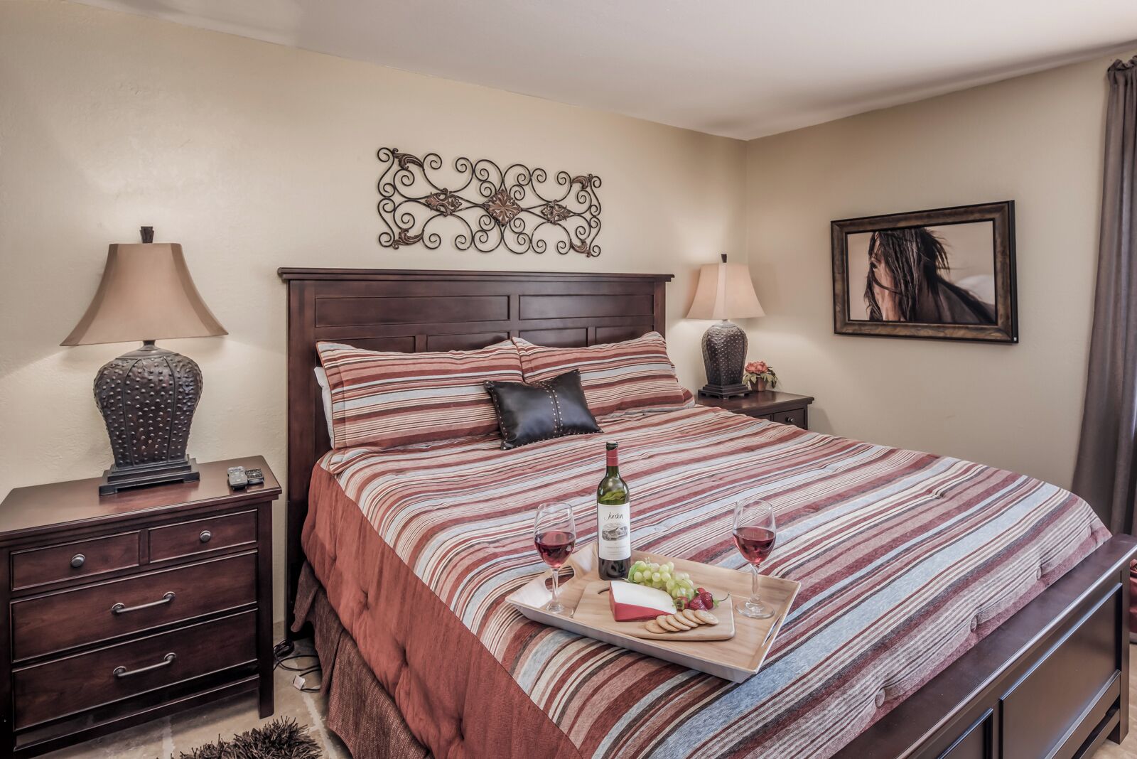 A neatly made bed with striped bedding, a bedside table with a lamp, and a tray with wine, grapes, and cheese.