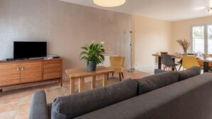 A modern living room with a flat-screen TV, a wooden cabinet, a grey sofa, a dining table with chairs, and a red pendant light. - Vacation home Lana by Interhome (Denneville)