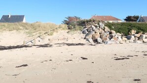 A sandy beach with a pile of rocks, a wooden staircase, and houses in the background. - Vacation home Lana by Interhome (Denneville)