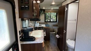 A compact RV interior with a kitchenette, a small bathroom, and a door leading to the outside.