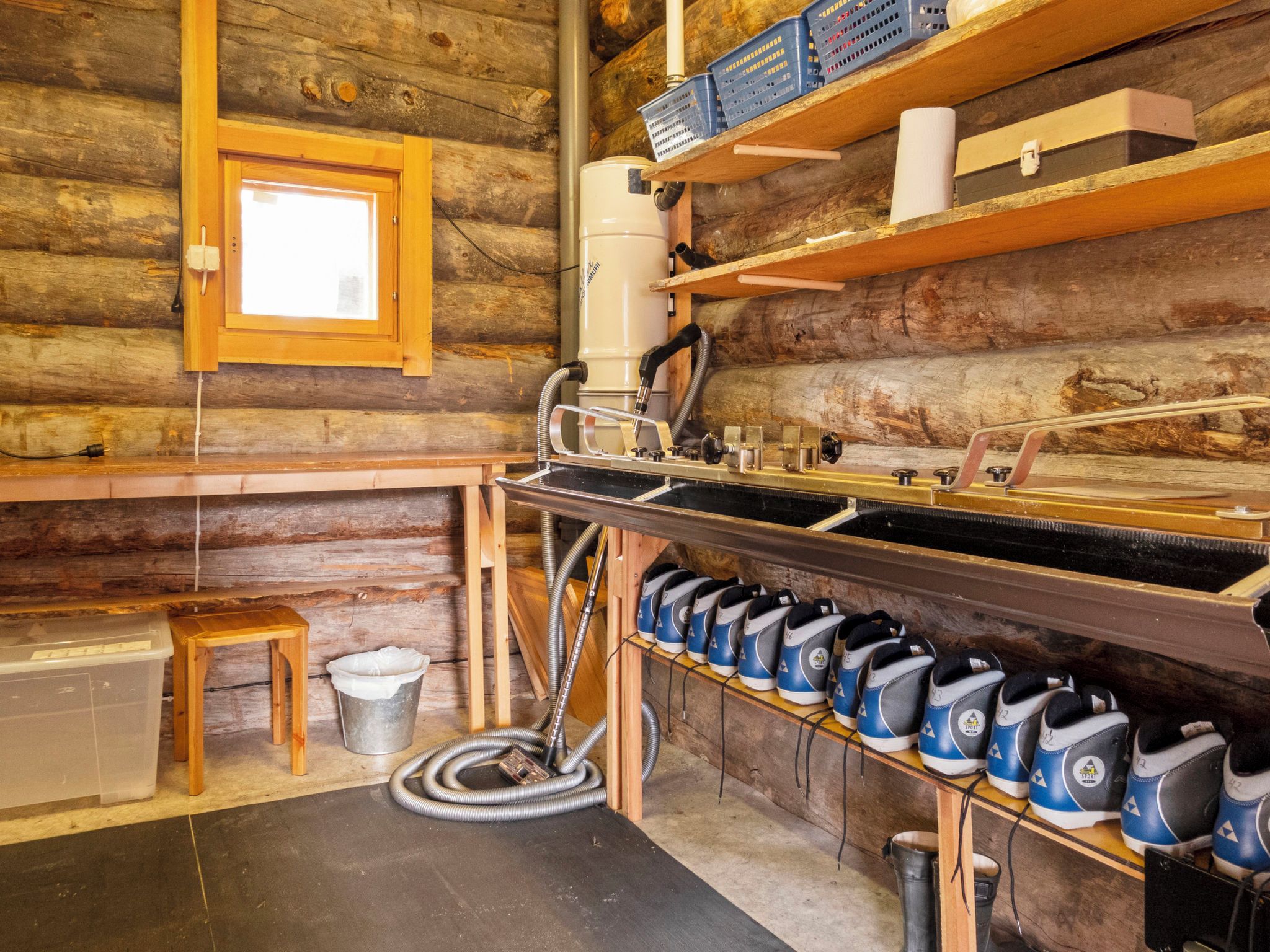 Interior rústico de una cabaña con un banco de madera, un lavamanos con mangueras, un cubo de basura y una fila de zapatos azules y blancos en un estante.