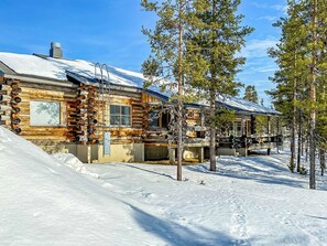 Una cabaña de troncos con techo nevado, rodeada de árboles y un paisaje invernal.