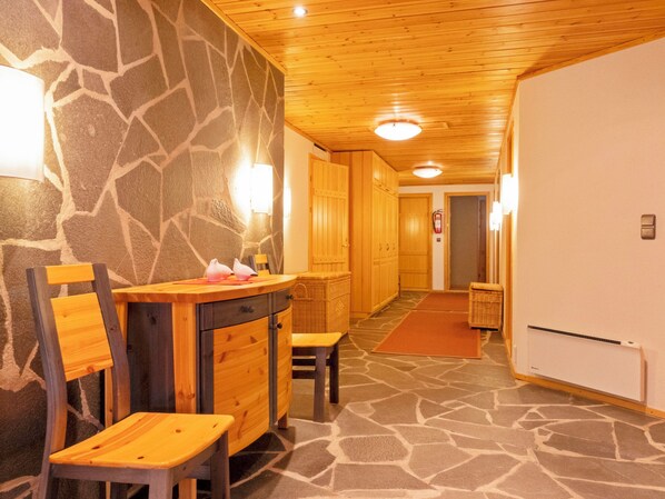 A hotel room with a stone wall, wooden ceiling, and a stone-patterned floor. There is a wooden dresser, a chair, and a small table with a bottle and a tissue box.