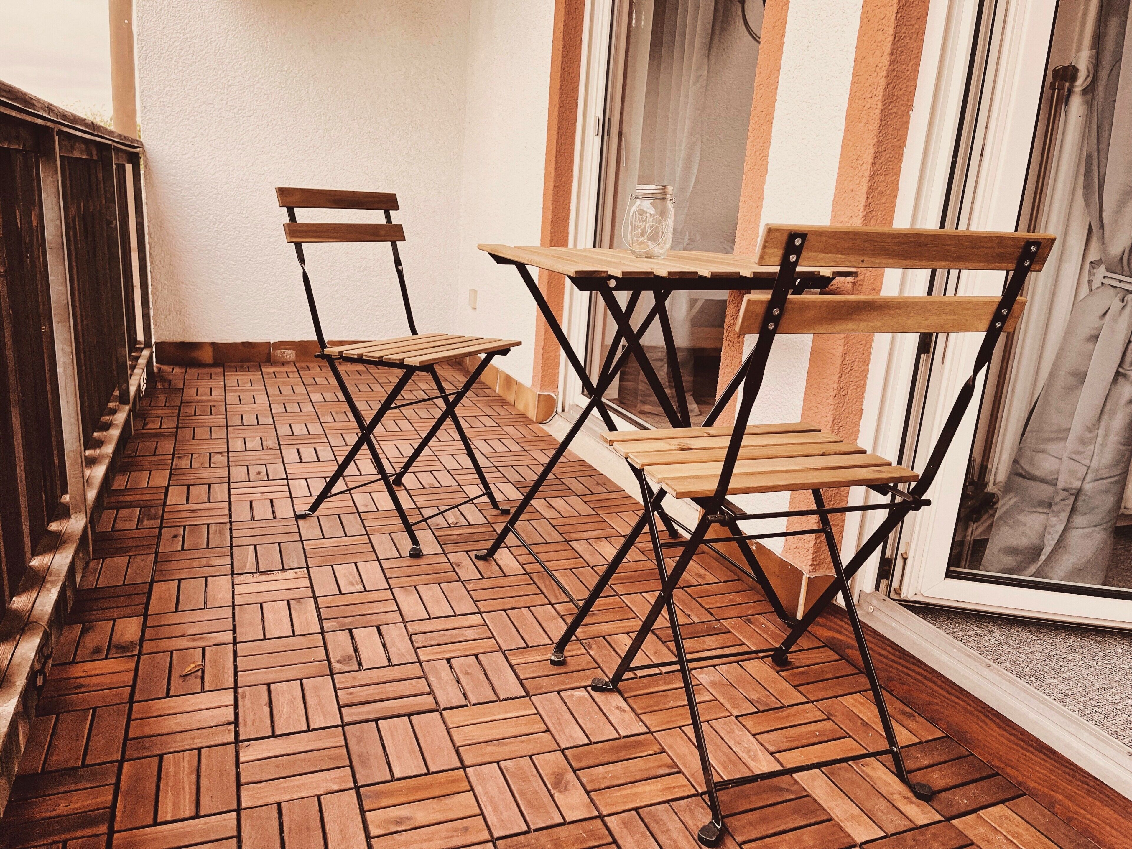 A small outdoor seating area with a wooden table and two chairs, a brick floor, and a window with curtains.