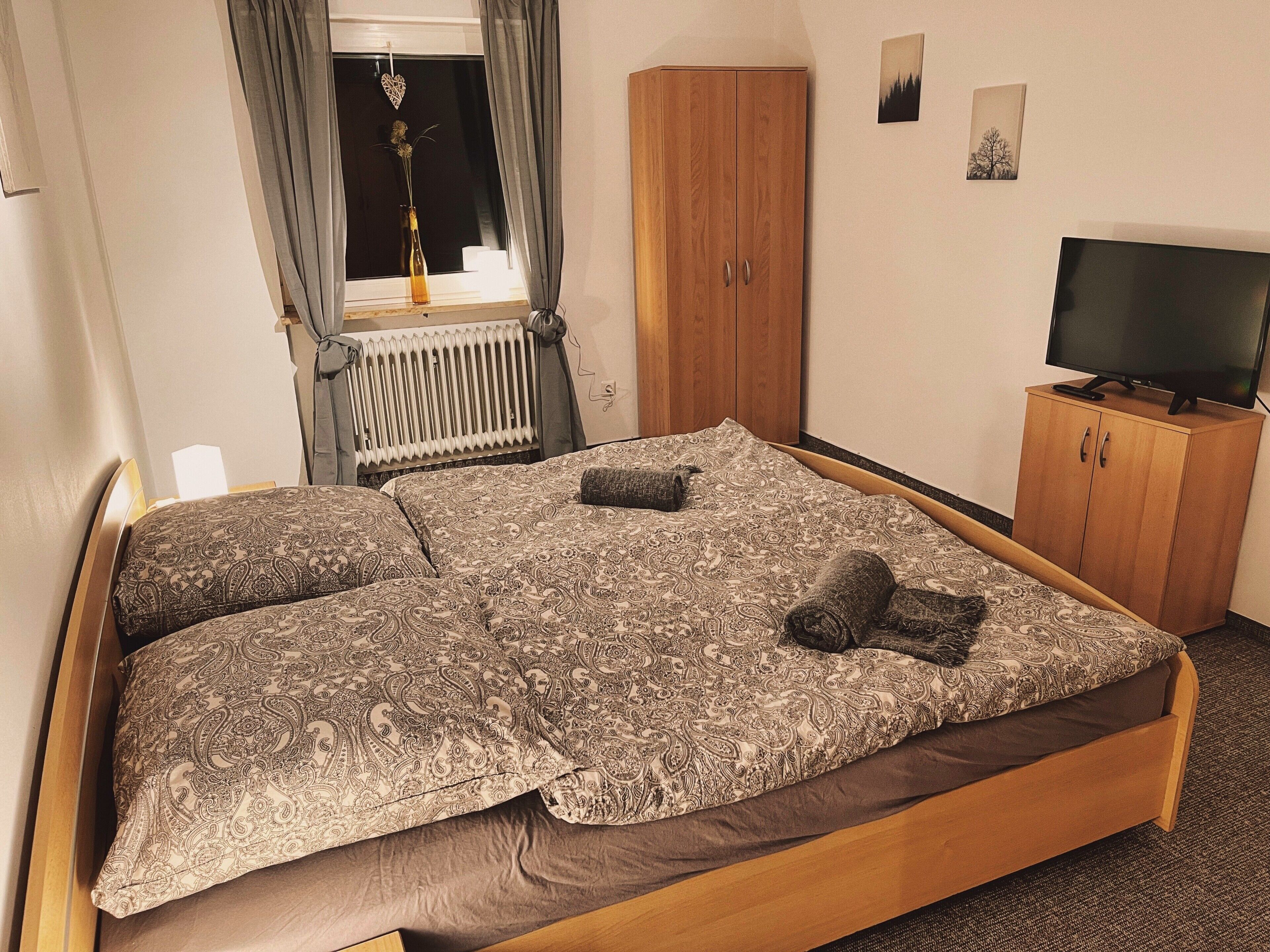 Ein Schlafzimmer mit einem Bett, einem Holzschrank, einem Heizkörper, einem Fernseher und einem Fenster mit Vorhängen.