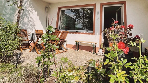 Un patio con sillas y una mesa de madera, una ventana con vistas a árboles y plantas en macetas.