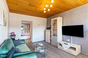 A modern living room with a green sofa, a glass coffee table, a flat-screen TV, and a wooden ceiling.