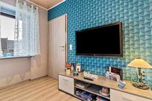 A room with a TV mounted on a textured blue wall, a wooden door, a desk with a lamp, and a window with a curtain.
