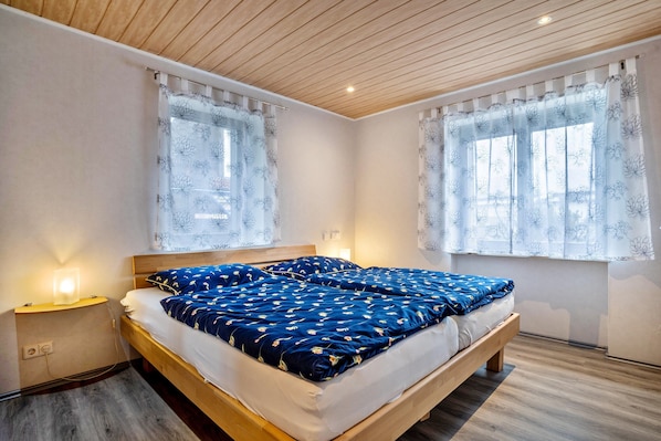 A bedroom with a wooden bed, a bedside table with a lamp, and two windows with floral curtains.