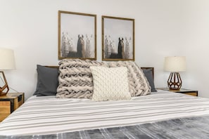 Cama bem arrumada com roupa de cama listrada, duas quadros na parede e duas luminárias de mesa.