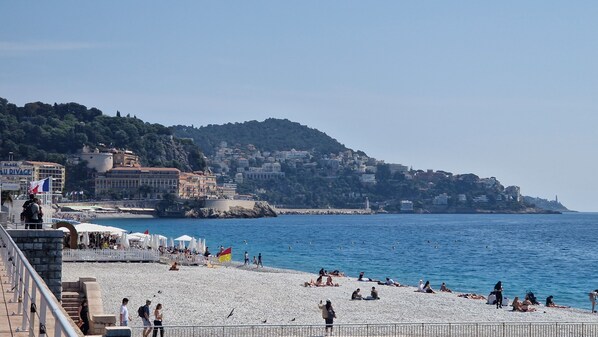 A coastal beach with people sunbathing and swimming, a promenade with umbrellas, and buildings on a hillside in the background.