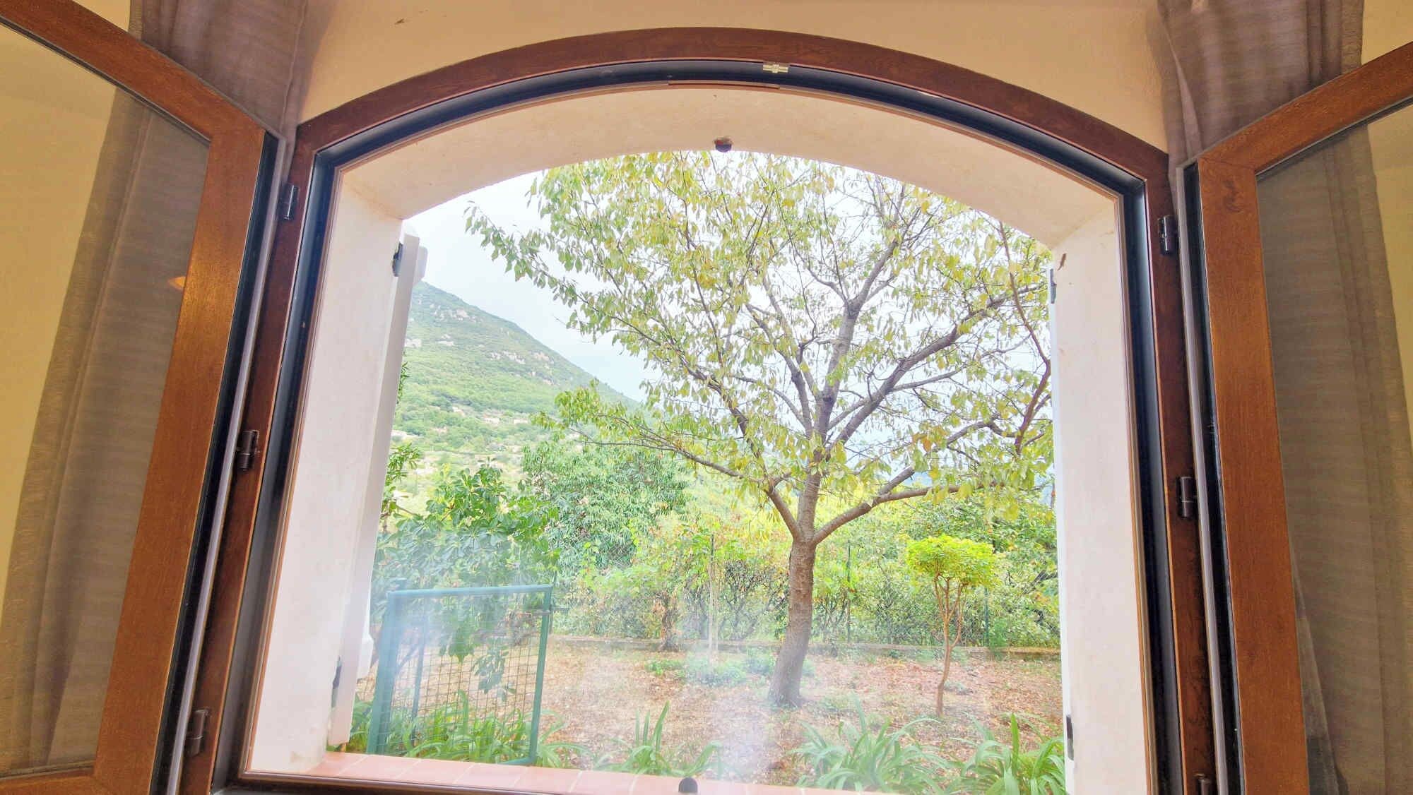 A room with a large arched window overlooking a garden with trees and a hilly landscape.