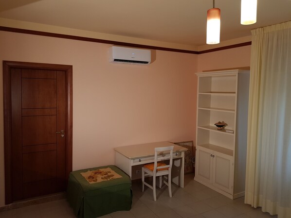 Une petite pièce avec une porte en bois, un bureau blanc avec une chaise, un pouf vert, une armoire blanche et un climatiseur fixé au plafond.