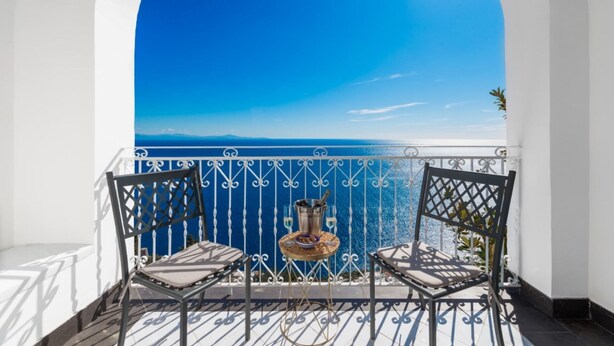 Ein Balkon mit zwei Stühlen und einem kleinen Tisch, mit Blick auf das klare, blaue Meer und ferne Berge.