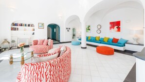 Ein geräumiger Wohnbereich mit weißer Einrichtung, eine Couch mit roten und weißen Kissen, eine blaue Couch mit bunten Kissen sowie ein roter Hocker.