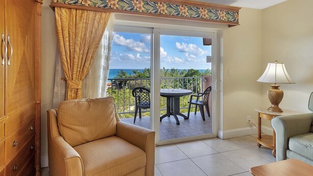 A room with a sliding glass door leading to a balcony with a view of the ocean. There is a beige armchair, a small round table with two chairs, and a lamp on a side table.