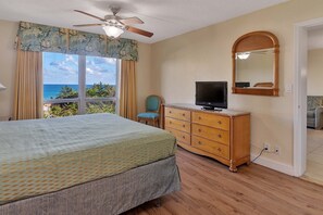 A bedroom with a bed, a dresser, a television, and a view of the ocean through large windows.