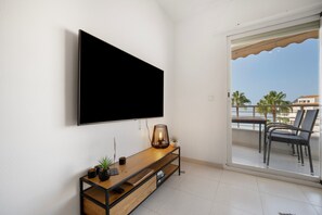 A modern living room with a large flat-screen TV mounted on the wall, a wooden console table with a lamp, and a view of the balcony with outdoor furniture.