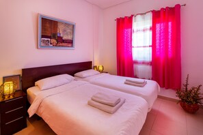 Habitación de hotel con dos camas, una mesita de noche con una lámpara, un cuadro en la pared y una ventana con cortinas rojas.