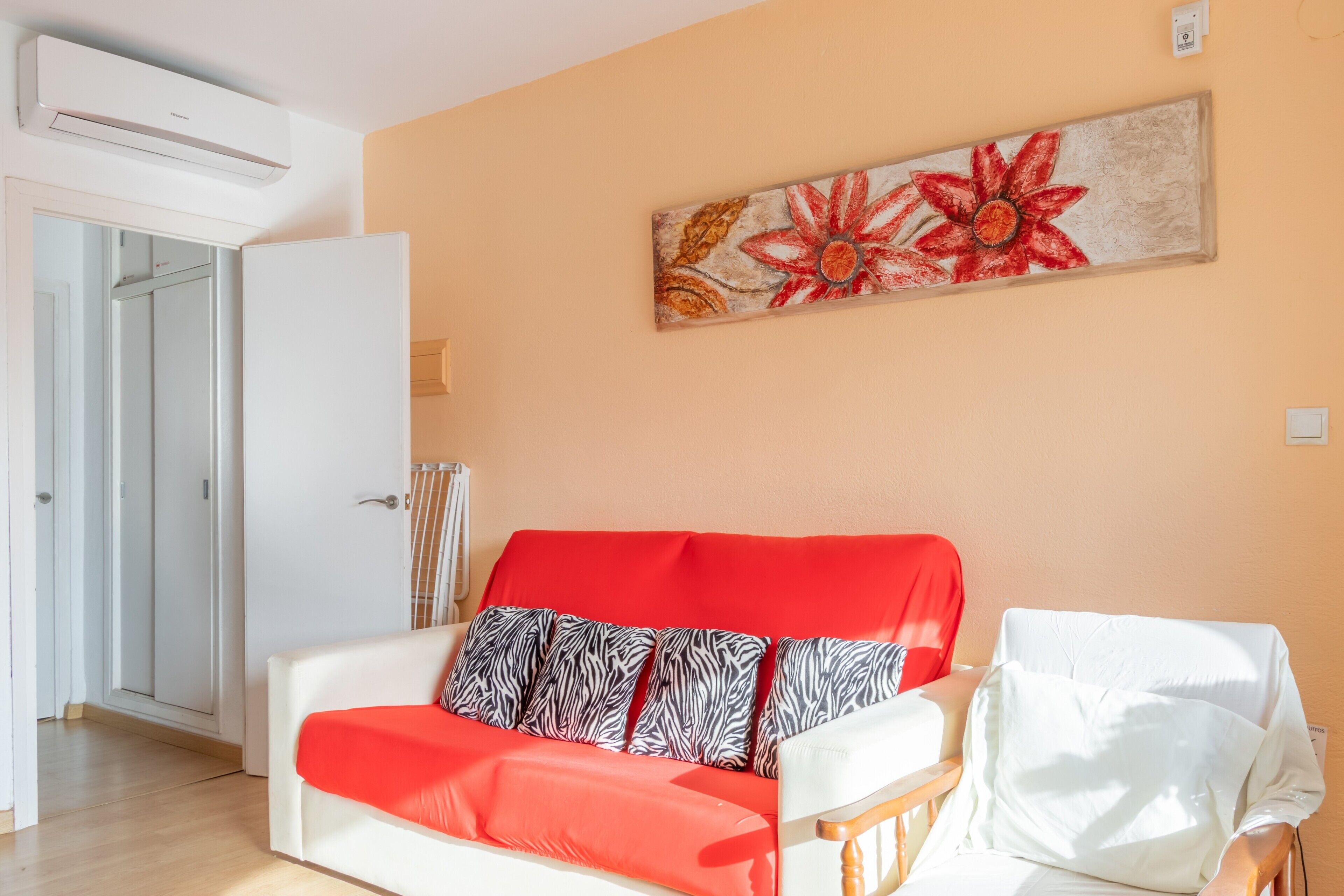 Een woonkamer met een rode en witte bank, een witte fauteuil, een schilderij aan de muur en een airconditioning.