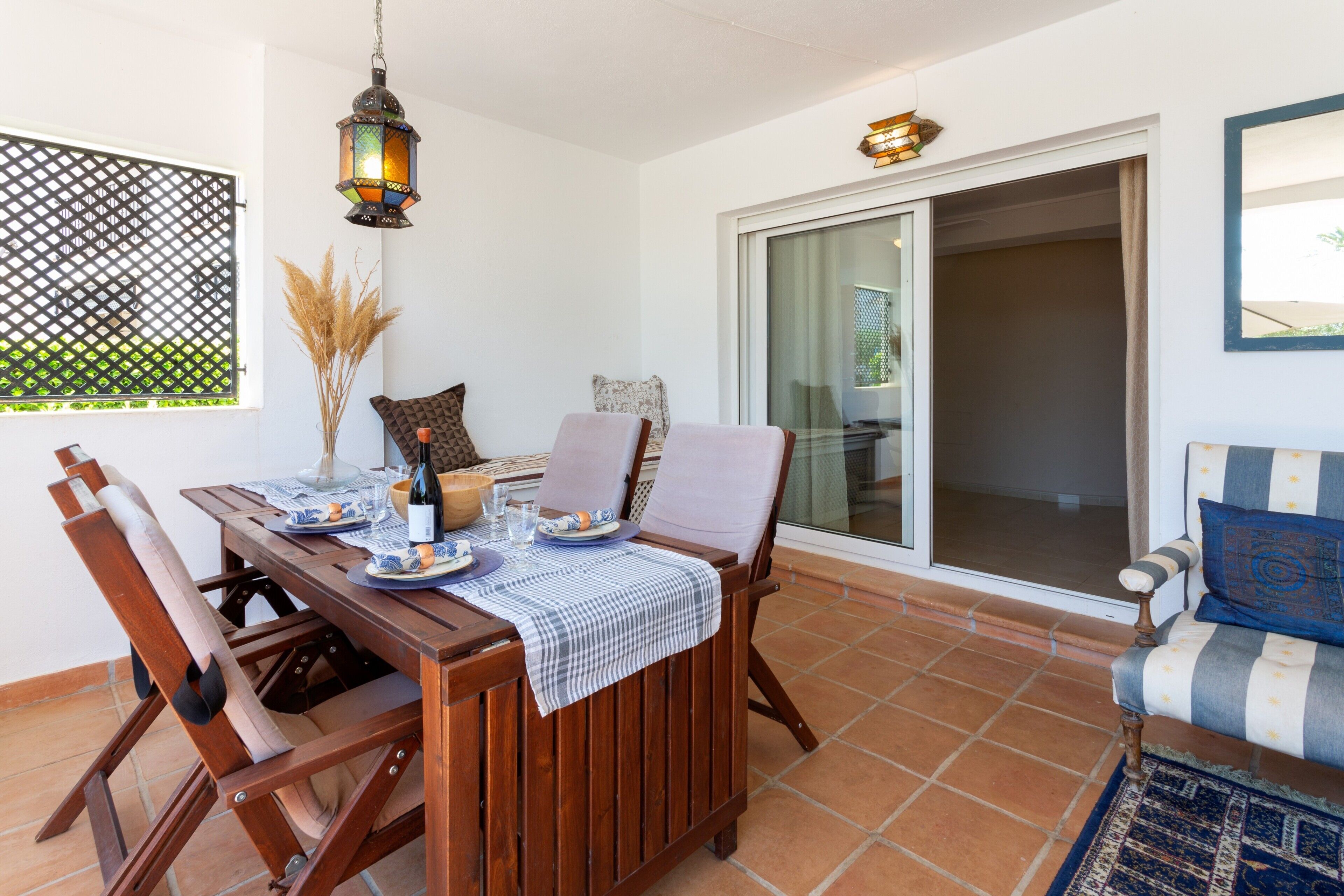 Eine Terrasse mit einem Holztisch, gedecktem Esstisch, gepolsterten Stühlen, einer hängenden Laterne und einer Glastür, die zu einem Außenbereich führt.