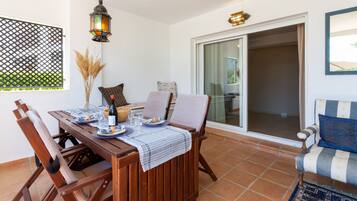 Eine Terrasse mit einem Holztisch, gedecktem Esstisch, gepolsterten Stühlen, einer hängenden Laterne und einer Glastür, die zu einem Außenbereich führt.