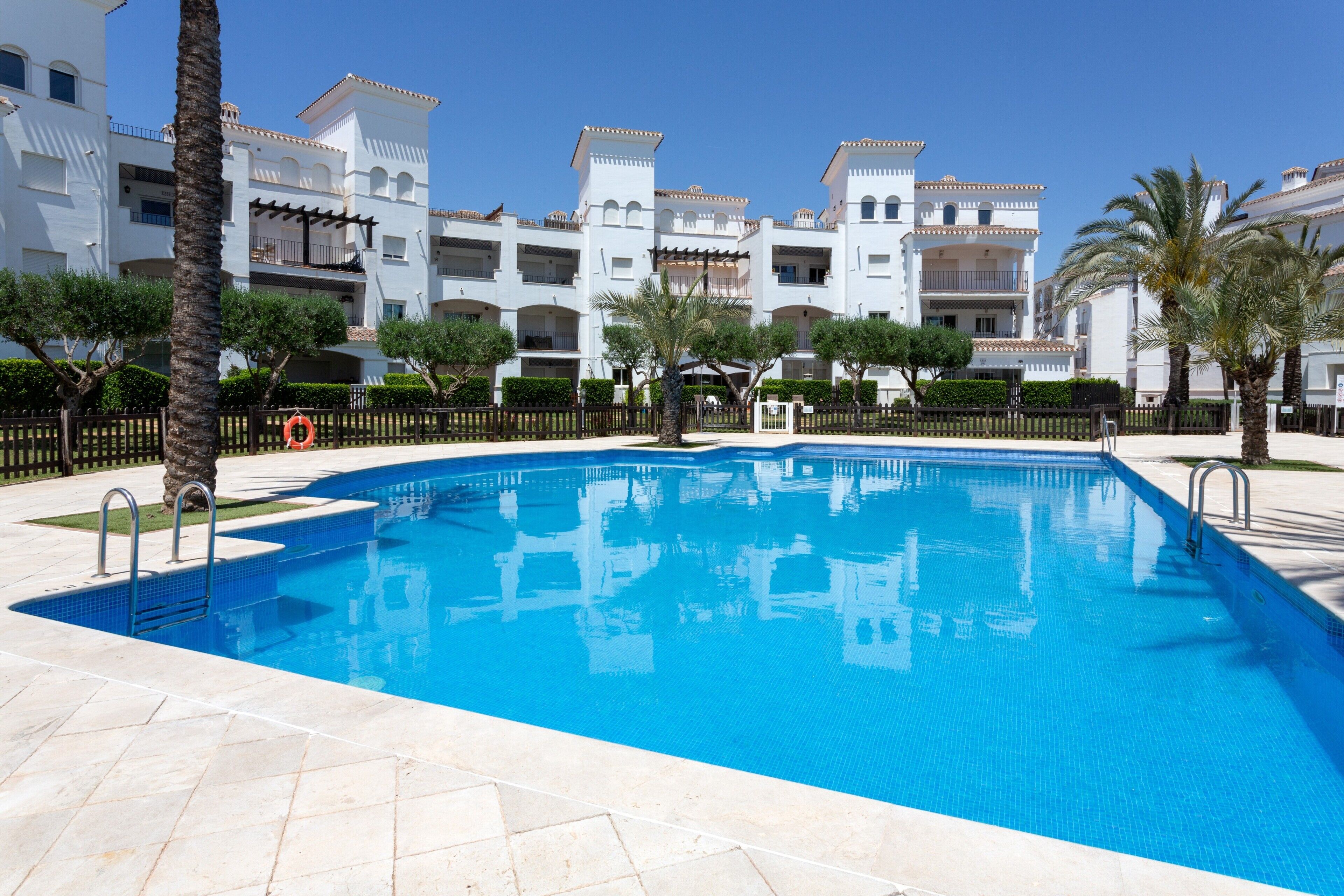 Une piscine à l’eau d’un bleu cristallin, entourée d’un bâtiment blanc doté de balcons et bordée de palmiers.