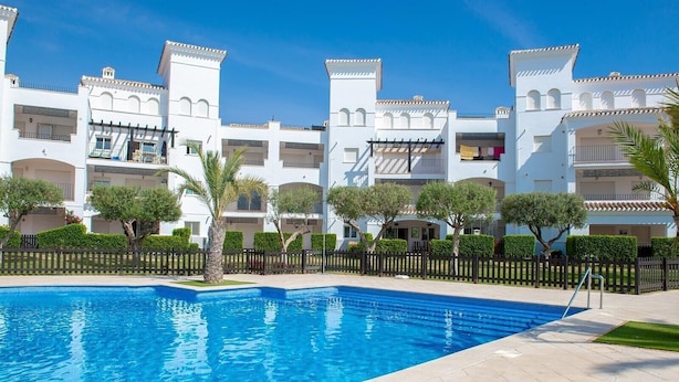 Una piscina con borde de baldosas, rodeada de edificios blancos con balcones y una valla.