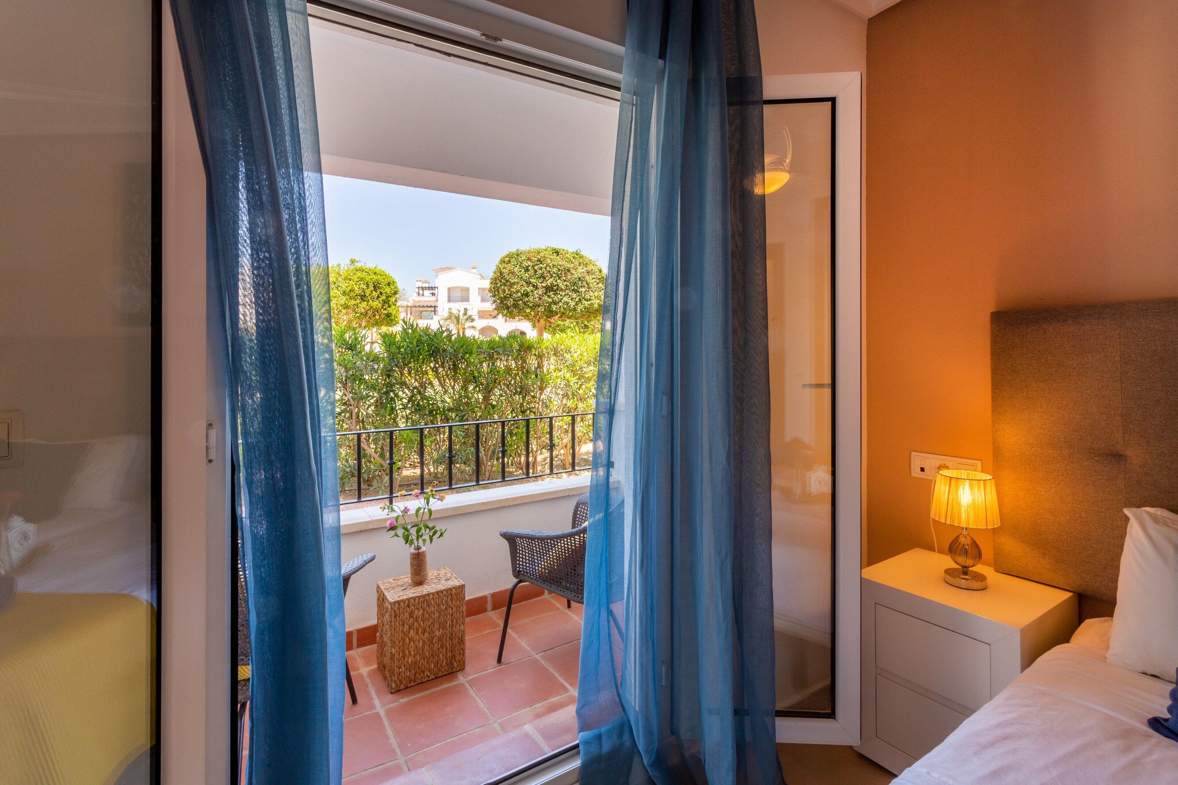 Una habitación de hotel con una cama, una mesita de noche con lámpara, una pequeña mesa con una planta, una silla y un balcón con vistas a zonas verdes y edificios.
