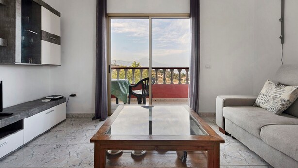 Een moderne woonkamer met een salontafel met glazen blad, een bank, een televisie en een balkon met een tafel en stoelen.