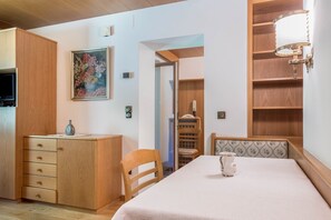 Una habitación con escritorio de madera, una silla, una cama con colcha blanca, una estantería con lámpara y un cuadro en la pared.