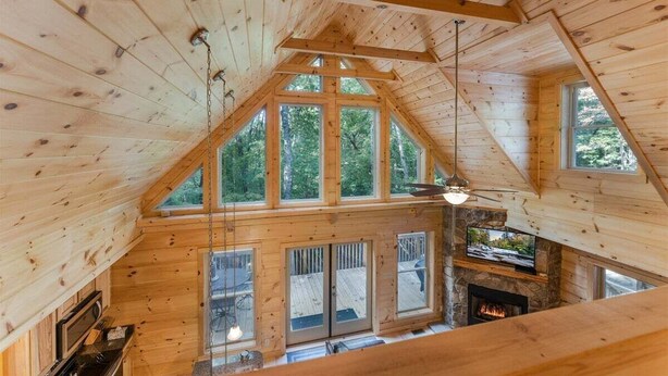 Interior de una cabaña de madera con chimenea de piedra, ventanas amplias y un televisor.