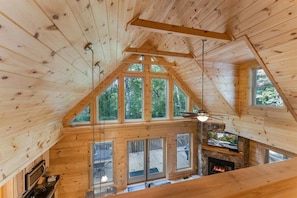 Interior de una cabaña de madera con chimenea de piedra, ventanas amplias y un televisor.