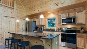 Interior de una cabaña de madera con cocina que incluye una isla grande, taburetes de bar, un microondas, una estufa y armarios de madera.