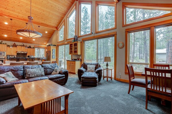 A spacious living area with a leather sofa, ottoman, and wooden coffee table. Large windows offer views of a forested area. - 5BR+Bunk,5.5BA,4 King,Near SHARC Pool,Hot tub,WiFi,A/C,Bikes,EV Chrg (Sunriver)