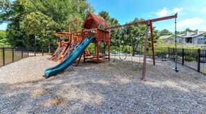 Children can play on the playground's slide and swings surrounded by trees and residential buildings.