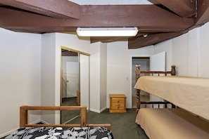 Una habitación pequeña y funcional con una cama alta, una mesita de noche de madera, un espejo y una puerta que da a otra habitación.