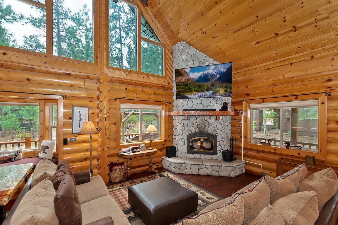 A log cabin interior with a stone fireplace, wooden walls, and large windows.