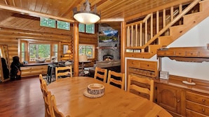 A wooden cabin interior with a dining area, fireplace, stone wall, and a staircase. - Red Cloud Cottage: Luxury Log Cabin Walking Distance to Snow Summit! Hot Tub! Game Room! Pet Friendly! EV Charger! (Big Bear Lake)