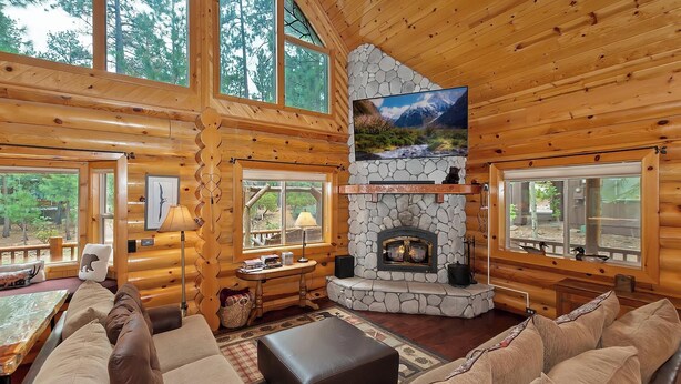 A cozy cabin interior with a stone fireplace, wooden walls, and large windows offering views of the forest. - Red Cloud Cottage: Luxury Log Cabin Walking Distance to Snow Summit! Hot Tub! Game Room! Pet Friendly! EV Charger! (Big Bear Lake)