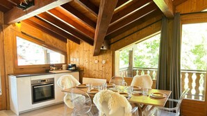 Una cabaña de madera con comedor que incluye una mesa y sillas de madera, una cocina con estufa y horno, y ventanas amplias con vistas al exterior.