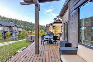 A modern outdoor deck with a dining area, a hot tub, and a view of a forested mountain. - Ski-In/Ski-Out Cabin with Private Hot Tub and Mountain Views (Big Sky)