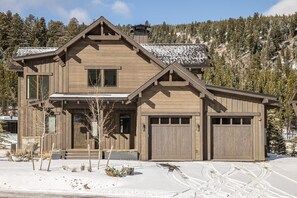 A two-story wooden cabin with a garage, surrounded by snow-covered ground and trees. - Ski-In/Ski-Out Cabin with Private Hot Tub and Mountain Views (Big Sky)