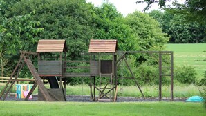 Une structure de jeu en bois, dotée d’une échelle et d’une balançoire, installée dans un espace verdoyant avec des arbres en arrière-plan.