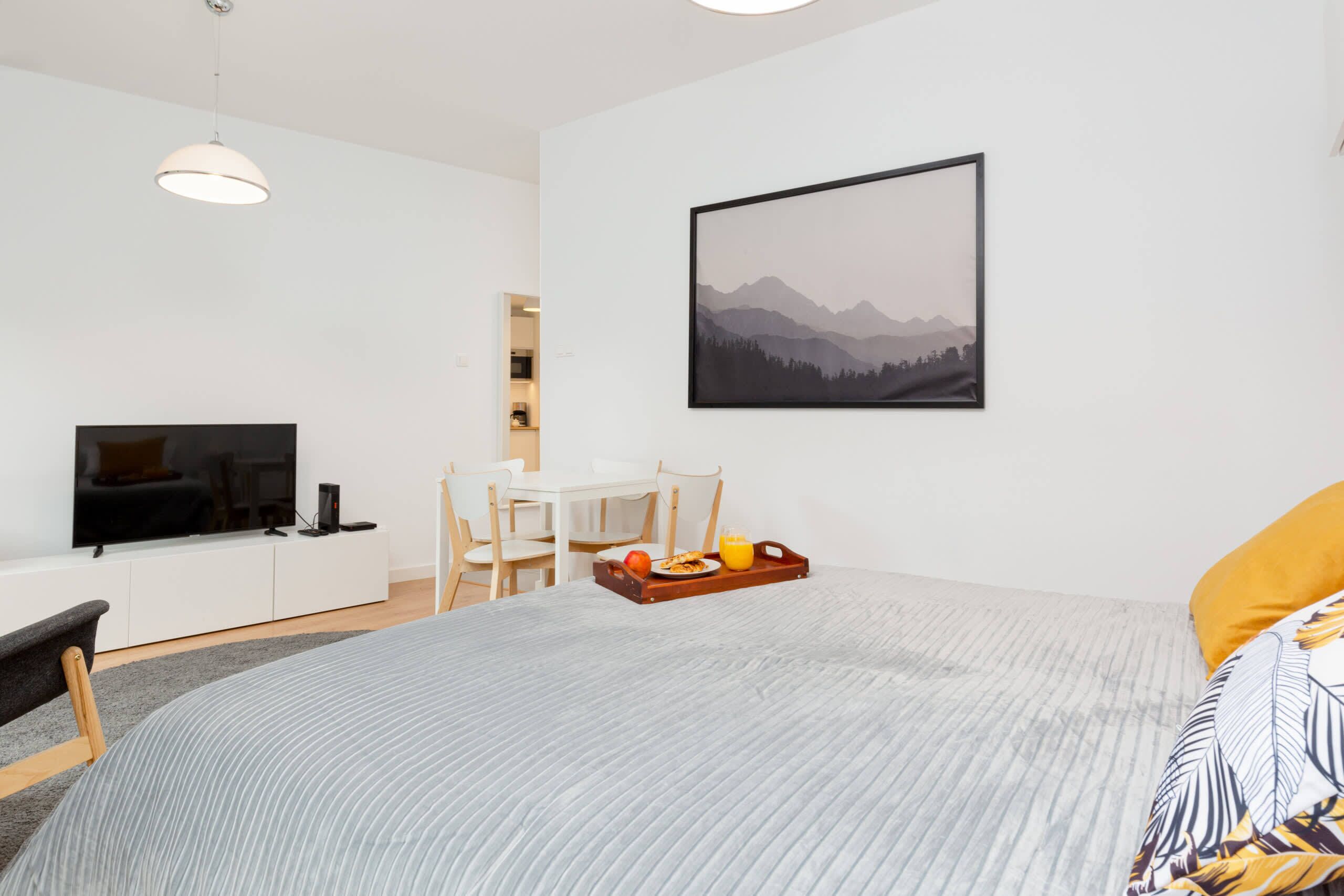 A modern hotel room with a bed, a flat-screen TV, a dining area with a table and chairs, and a framed landscape painting on the wall.